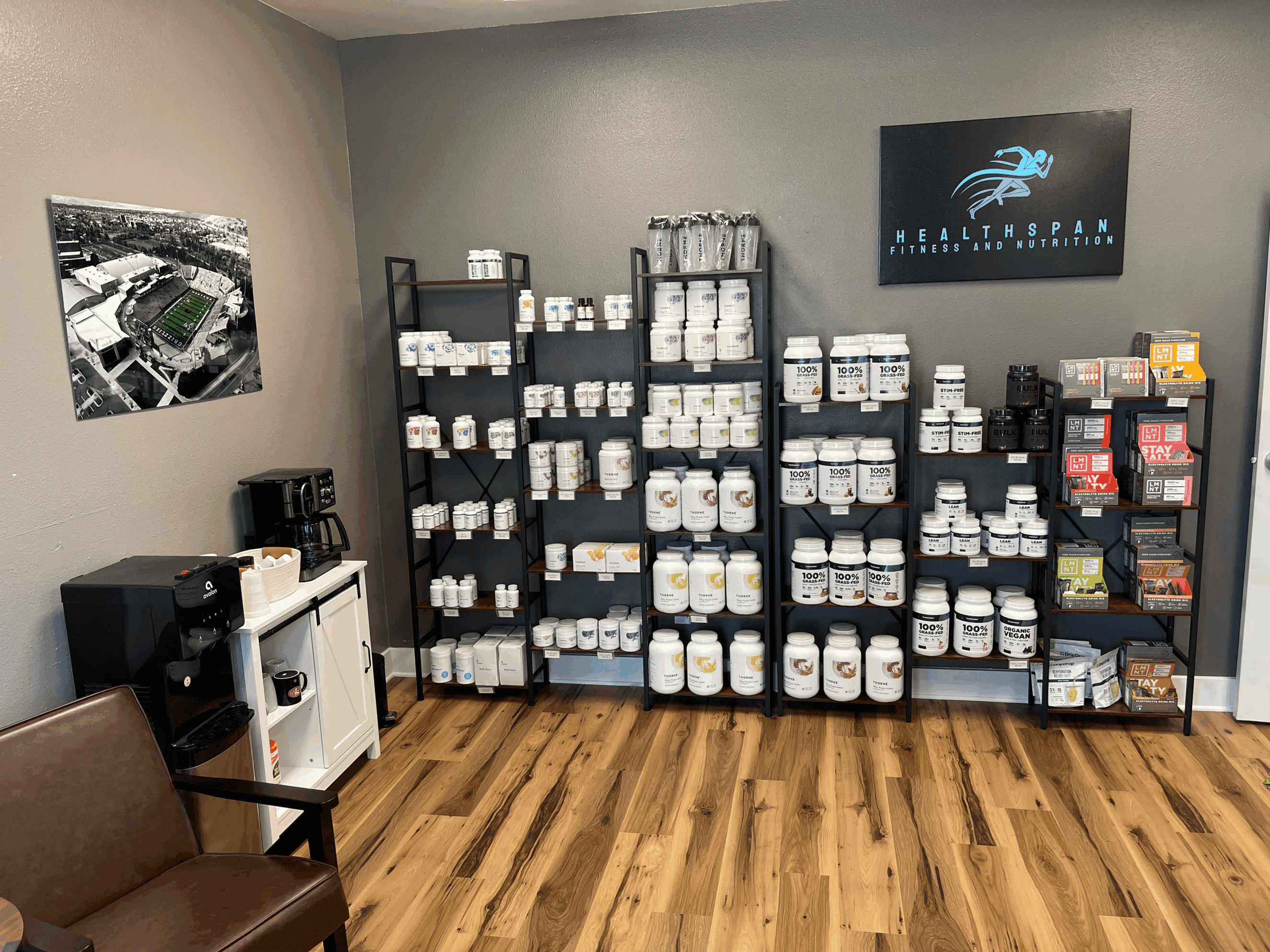 5928a6_5291c940190045aea40c9a624afb968e~mv2 shelves stocked with premium supplements and nutraceuticals including protein powders, vitamins, and energy products at Healthspan Fitness and Nutrition center with a coffee station and a framed aerial photo on the wall