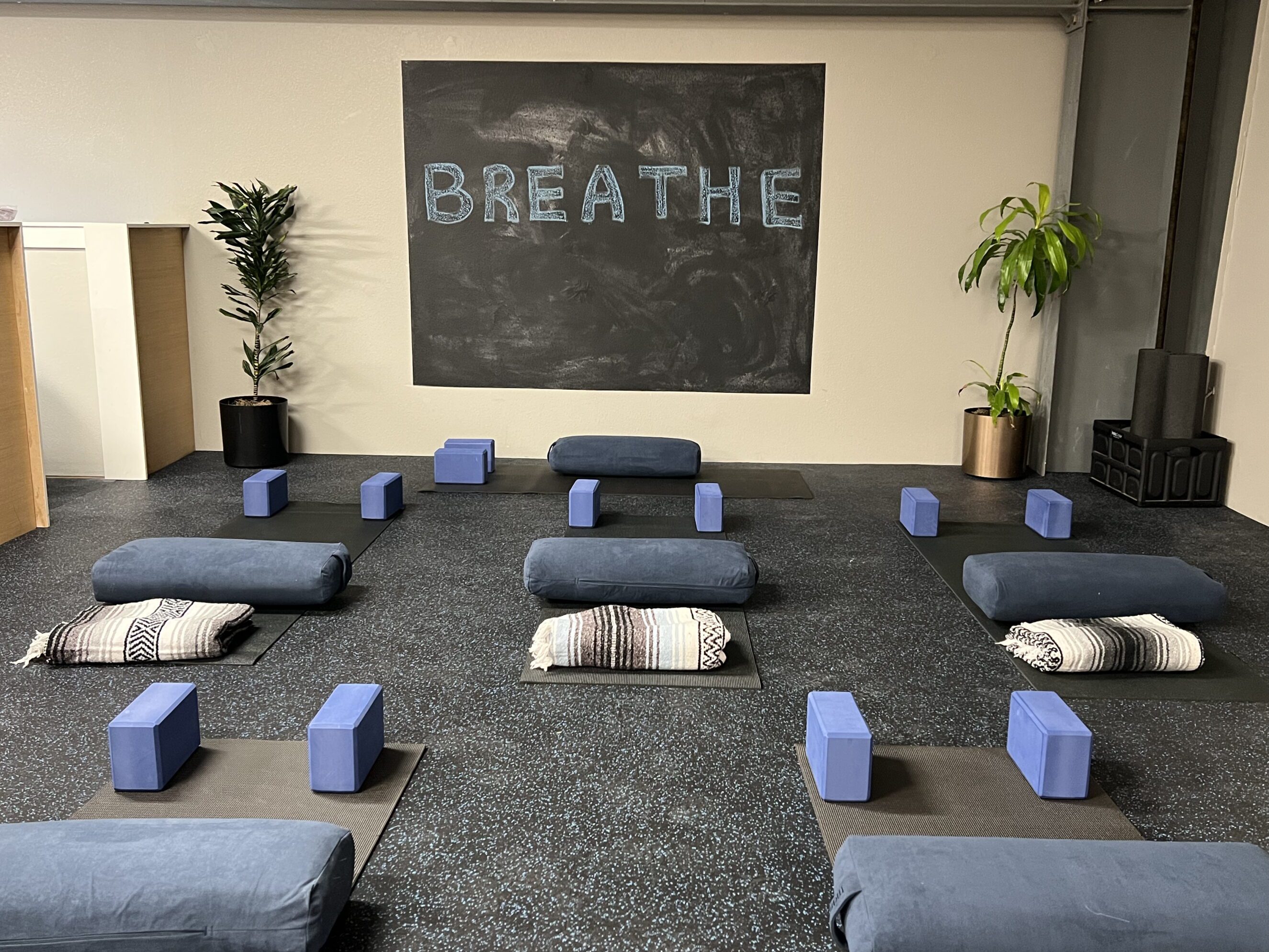 5928a6_6481e95cdac6458890fc2e8d21de1b53~mv2 Yoga and restorative movement studio with neatly arranged mats, bolsters, blankets, and yoga blocks under a chalkboard reading "BREATHE," promoting mind-body balance and stress relief as part of holistic wellness services.