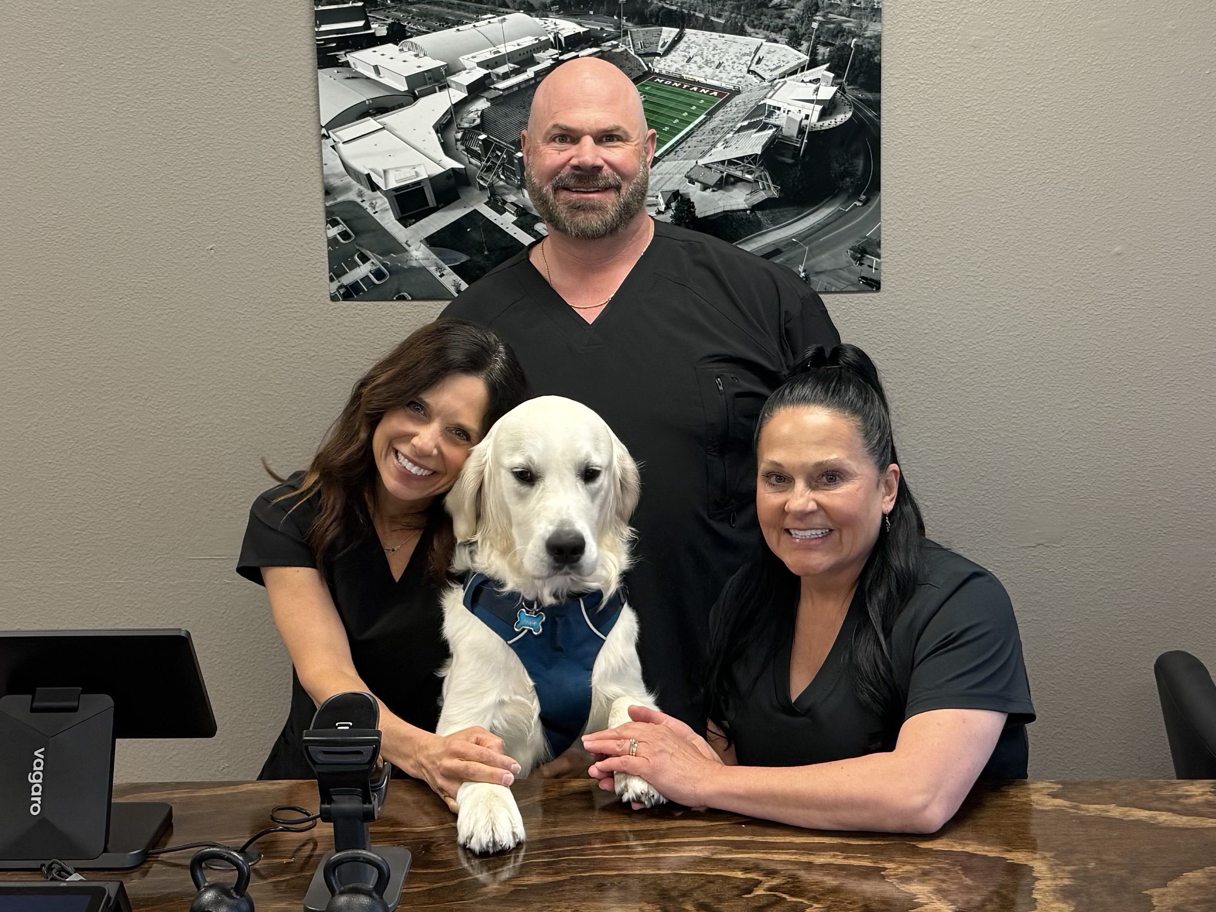 groupphotos Three Healthspan Medical team members in black scrubs, smiling around a golden retriever wearing a blue vest, seated at a wooden desk with medical and fitness equipment, with a black-and-white aerial photo of Montana stadium on the wall behind them.